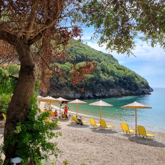 חוף קסום ונסתר ליד פארגה (Parga) ביוון - מפרץ טורקיז צלול עם מיטות שיזוף צהובות ושמשיות קש בלב הטבע