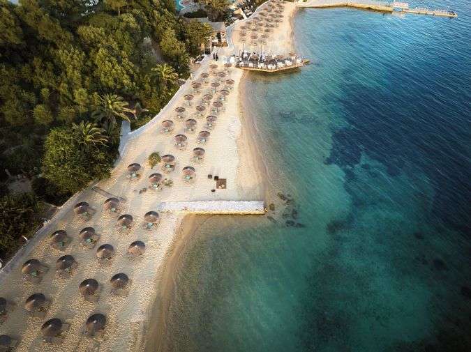 מבט אווירי על חוף הים הזהוב והמים הצלולים של ריזורט Eagles Palace בחלקידיקי, עם שמשיות קש הפרוסות לאורך החוף וצמחייה ירוקה עשירה