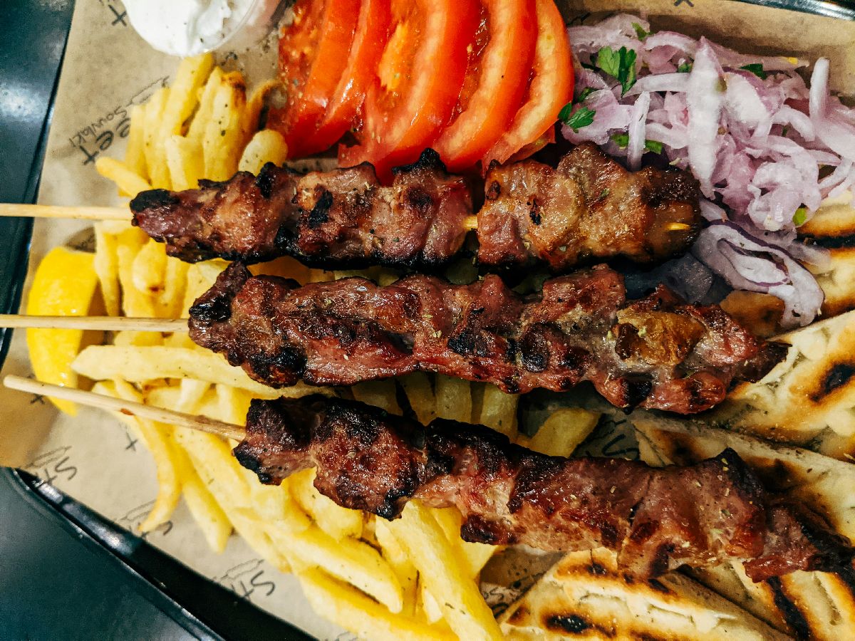 מגש הגשה עם שלושה שיפודי בשר בגריל לצד צ'יפס, פרוסות עגבנייה, בצל סגול קצוץ ופיתה יוונית